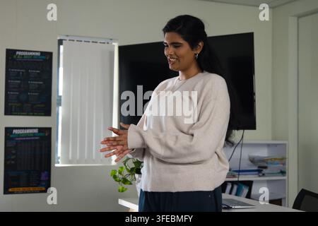 Indian woman wearing beige sweater gesturing while presenting in office with laptop and monitor Stock Photo