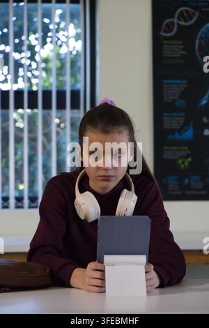 Adolescent female student studying at class desk in maroon hoodie, wearing headset and using tablet Stock Photo