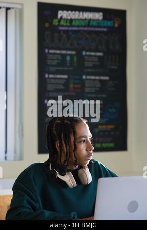 African American teen sitting at desk wearing headphones, using laptop, code poster behind, blinds Stock Photo