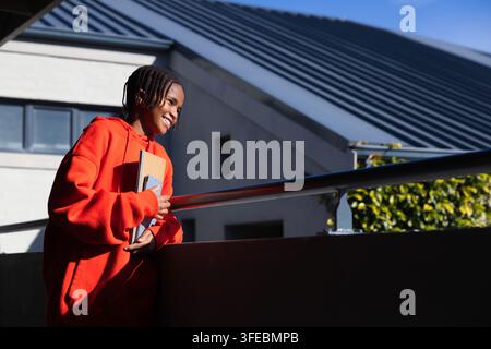 African male youth standing on balcony holding notebooks, wearing red hoodie by railing, copy space Stock Photo
