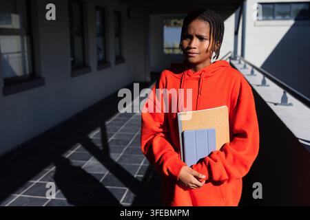 African youth male in red hoodie holding notebook, tablet on school walkway by windows, copy space Stock Photo