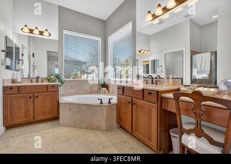 Spacious master bathroom interior featuring double wooden vanities, beige marble countertops large mirrors soft lighting and corner bathtub with glass Stock Photo