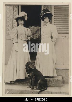 Two women, each wearing long dresses and hats, stand on steps with a dark-colored dog between them, in front of a doorway with patterned curtains and a shuttered window. Stock Photo