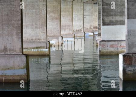 Perspective from underneath a large modern concrete bridge, where massive rectangular piers recede into calm water, forming repeating rows and depth. Stock Photo