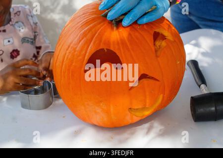 A large orange pumpkin being carved into a jack-o'-lantern for Halloween using cookie cutters and a mallet on a table. Stock Photo