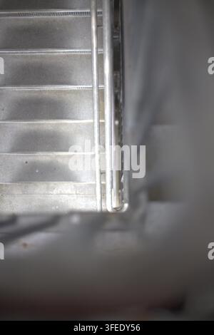 A high-angle view looking down a metal staircase with steel handrails and blurred foreground framing in an industrial building. Stock Photo