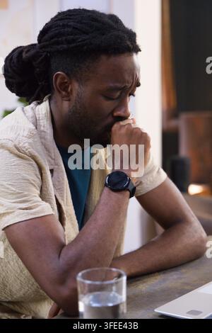 Adult African man in beige shirt resting elbow holding hand-to-mouth while using laptop at table Stock Photo