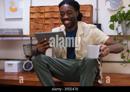 Man sitting on bench in home studio, wearing beige shirt, holding tablet, white mug Stock Photo