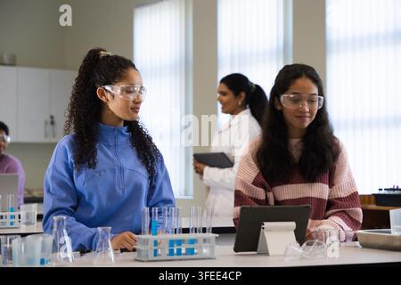 Two teenage female students in pullover goggles consulting tablet observing test tubes at lab bench Stock Photo