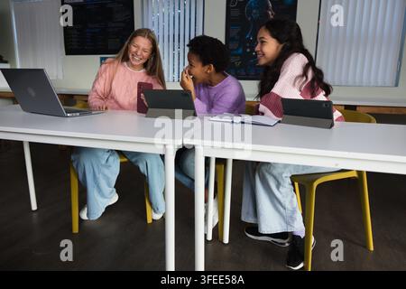 Diverse teenage female friends laughing sharing tablet and phone while using laptop in classroom Stock Photo