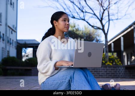 Asian female child sitting on campus plaza, using laptop with purple headphones, wearing sweater Stock Photo