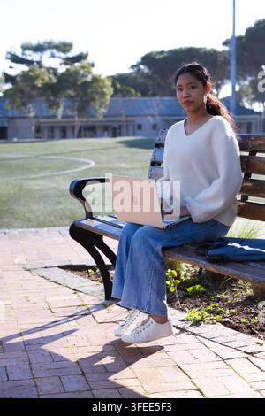 Asian child girl sitting on wooden park bench working on laptop beside backpack, copy space Stock Photo