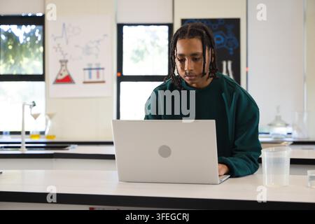 African American youth in green sweater working on laptop at lab with measuring cup and flasks Stock Photo