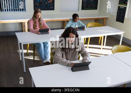 Diverse youth classmates studying on tablets with headphones at classroom with blinds and posters Stock Photo