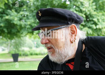 Older man wearing a black captain's hat with an anchor emblem, glasses, and multiple earrings. He has a white beard and is dressed in black clothing. Stock Photo