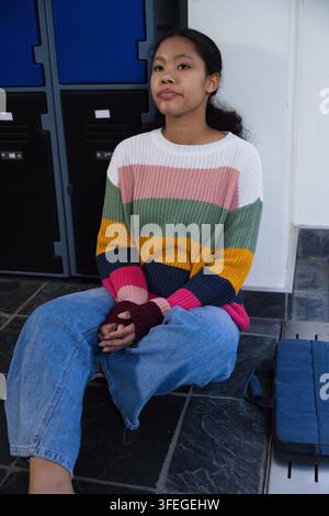 Asian child sitting in striped sweater and jeans on corridor floor near lockers and navy bag Stock Photo