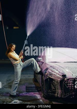 Happy smiling woman spraying pink active foam on luxury car using high pressure washer wand at outdoors self-service station at night time. Female dri Stock Photo