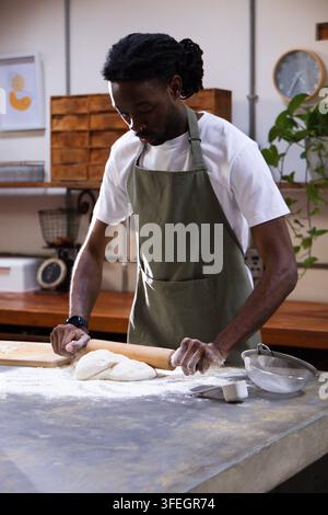 Adult African American man in olive apron using wooden rolling pin rolling dough on floured counter Stock Photo