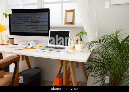 Software development and remote work burnout, messy home office desk with computer and laptop showing programming code in a bright sunlit workspace Stock Photo