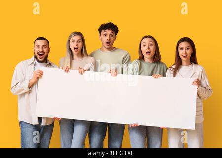 Marketing announcement and promotion, diverse group of surprised young people holding a large blank white sign for copy space on a yellow background Stock Photo