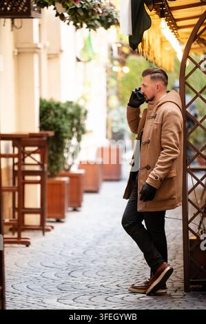 Stylish man enjoys a warm beverage while strolling through a charming outdoor cafe in autumn, surrounded by lush greenery and cozy decor Stock Photo