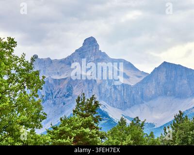 Dramatic Rocky Mountain Peak Over Forested Alpine Landscape Under Cloudy Sky Stock Photo