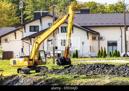 Yellow tracked excavator works on land development, removing dark silt from a canal bank with modern suburban homes in background. Stock Photo