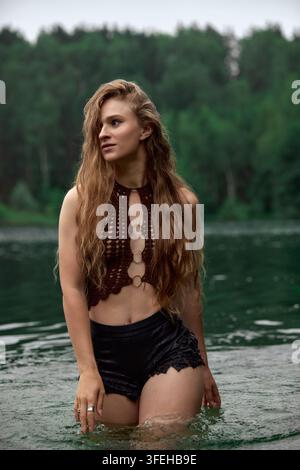 Pensive young woman in boho crochet top stands waist deep in serene lake water. Natural diffused light. Moody green forest background creates connecti Stock Photo