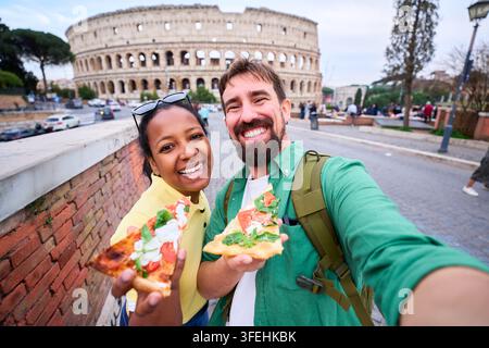 Happy couple taking a fun selfie while eating pizza in front of colosseum in Rome. Stock Photo