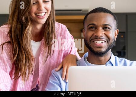 A happy young couple working together at a laptop, smiling and collaborating in a bright home office, sharing ideas and enjoying teamwork Stock Photo