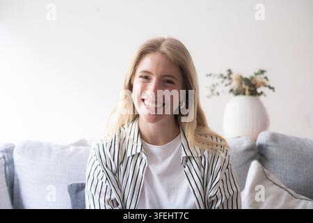 Young smiling woman sitting on a couch in a bright modern living room, casual striped shirt and white tee, relaxed portrait conveying warmth, friendli Stock Photo