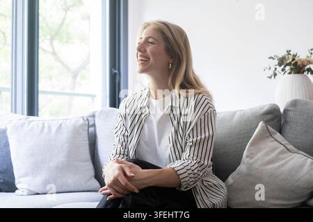 Smiling woman relaxing on a modern living room sofa in natural daylight, wearing a striped shirt and white tee, looking content and comfortable in a b Stock Photo
