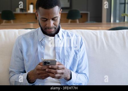 Young man sitting comfortably on a light sofa, engrossed in his mobile phone, communicating or browsing with focused concentration while enjoying casu Stock Photo