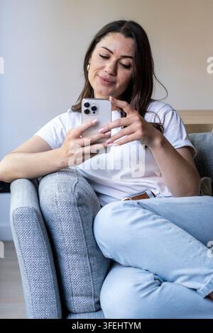 Young woman sitting on a couch at home using a smartphone, relaxed and smiling while scrolling social media and texting in a casual outfit. Stock Photo