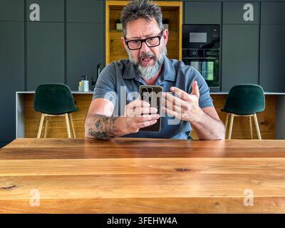 Middle-aged man with glasses and beard using smartphone for a video call in a modern kitchen, expressive and engaged during a remote conversation, ill Stock Photo