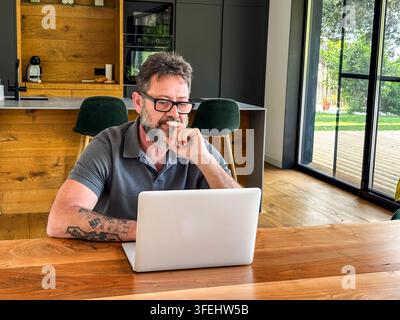 Middle-aged bearded man with glasses concentrated on laptop at wooden table in modern home, working remotely with garden view, focused on planning and Stock Photo