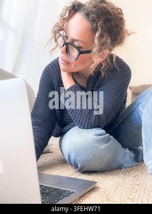 A woman in a cozy knit sweater and jeans works on a laptop from home, wearing glasses and sitting on the floor, conveying a relaxed remote work and st Stock Photo