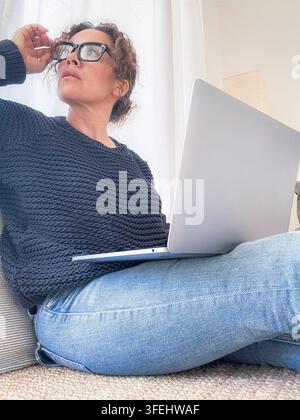 A casually dressed woman works remotely on a laptop at home, wearing a knit sweater and jeans. She sits on the floor with glasses, looking thoughtful Stock Photo