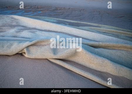 A detailed close-up shot capturing folded layers of a heavy, light-colored fine mesh fishing net resting on wet beach sand. Soft twilight lighting hig Stock Photo