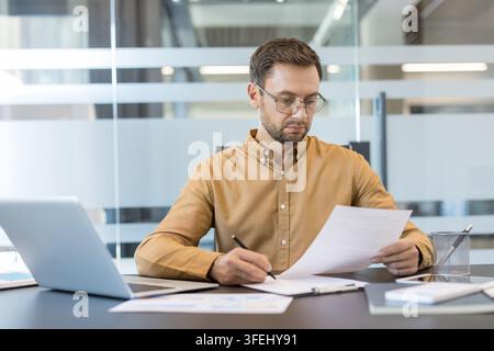 Businessman in glasses analyzing important documents, reviewing reports, and writing notes with a pen while sitting focused at a modern office desk with a laptop Stock Photo