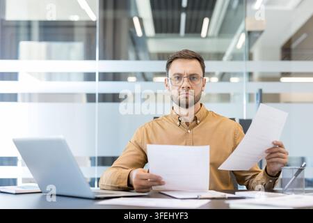 Businessman in glasses holding and reviewing paper documents at a desk with a laptop, reflecting focus and concentration on important paperwork and financial data Stock Photo