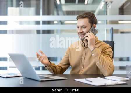 Happy businessman having a phone conversation while simultaneously working on his laptop at a modern office desk, managing tasks and communicating effectively Stock Photo
