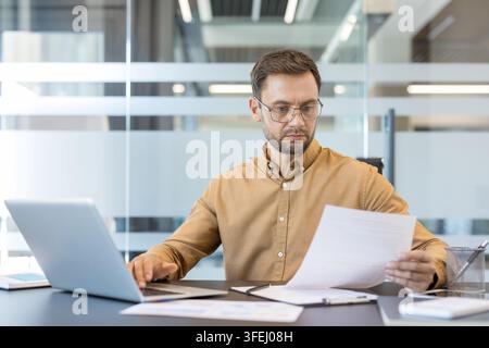 Concentrated man with glasses working on a laptop and reviewing financial papers at his desk in a modern corporate office, embodying diligence and business analysis Stock Photo