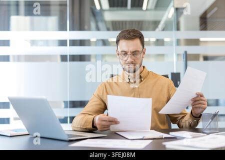 Businessman wearing eyeglasses sitting at an office desk, thoroughly examining several financial documents and paperwork while working on business analytics and accounting tasks Stock Photo