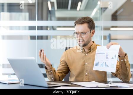 Businessman working in a modern office. Showing a document with graphs and charts. Feeling stressed and confused while analyzing financial data on his laptop. Indicating problems or unexpected results Stock Photo