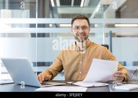 Young businessman smiling while managing corporate paperwork and using computer technology at a modern office desk, embodying success, productivity, and a positive work environment Stock Photo