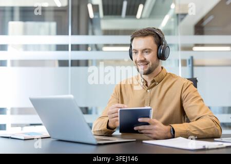 Young man wearing headphones sitting at a desk in a modern office, using a laptop for an online meeting, and taking notes in a notebook, smiling and focused Stock Photo