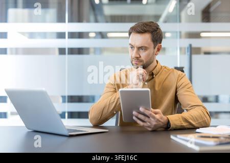 Young businessman concentrating while comparing information on a digital tablet and laptop, reflecting on business strategy and problem-solving in a modern office environment Stock Photo