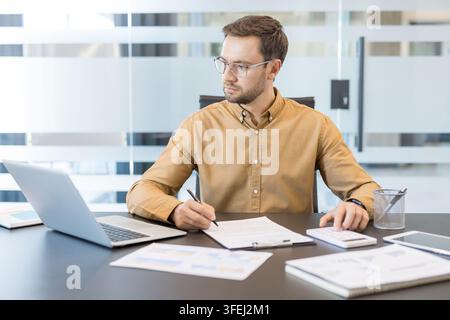 Young businessman focused on financial calculations and data analysis, writing notes and using a calculator while working at his desk in a modern office environment Stock Photo