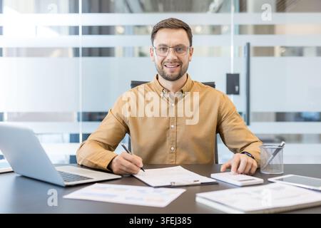 Smiling young businessman sitting at desk, reviewing documents, calculating finance with a calculator, and working on a laptop, representing business management and accounting Stock Photo